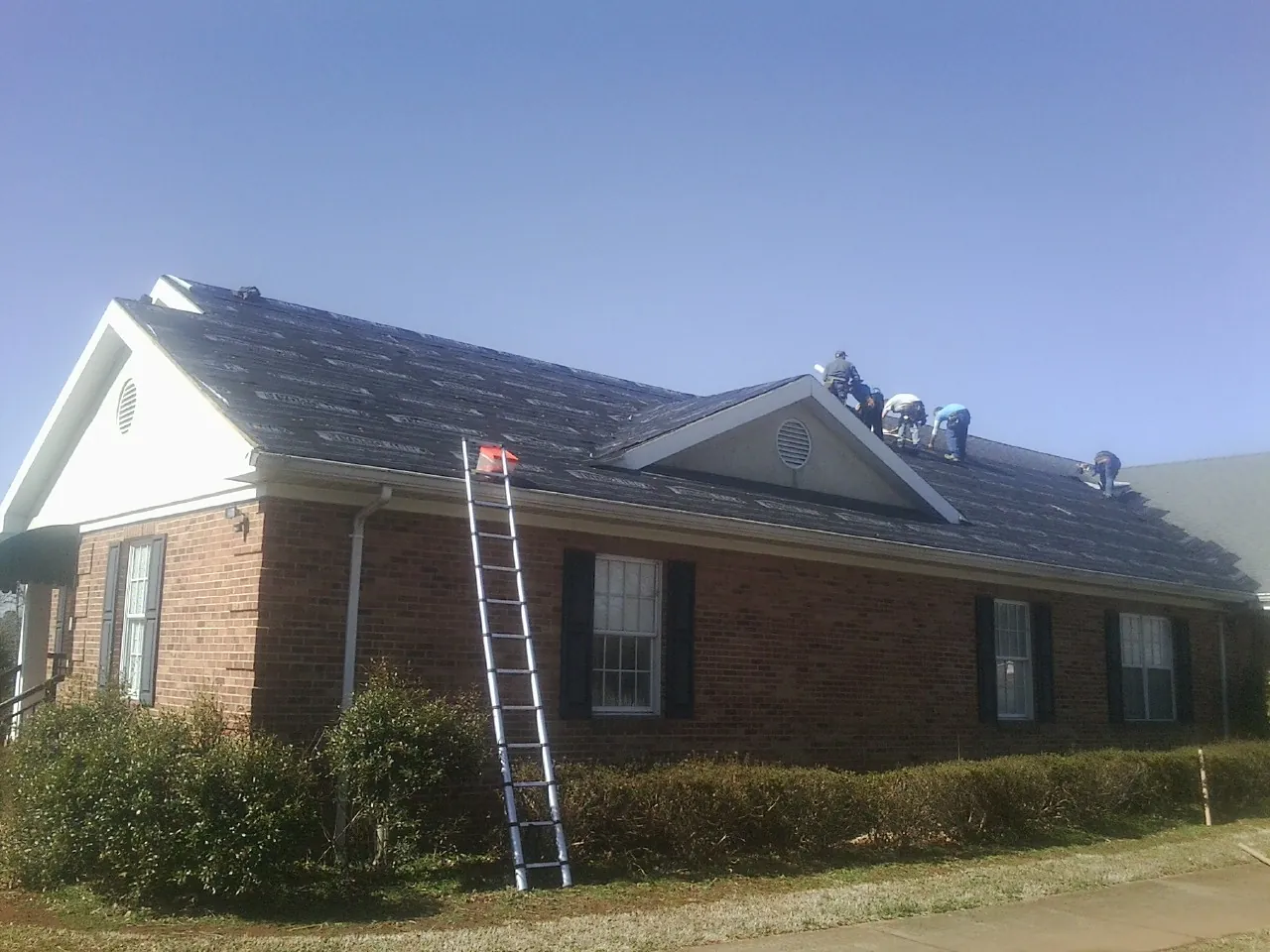 Crew tearing shingles off a brick ranch roof with a ladder set — Emergency Roof Repair work by our crew in Foley